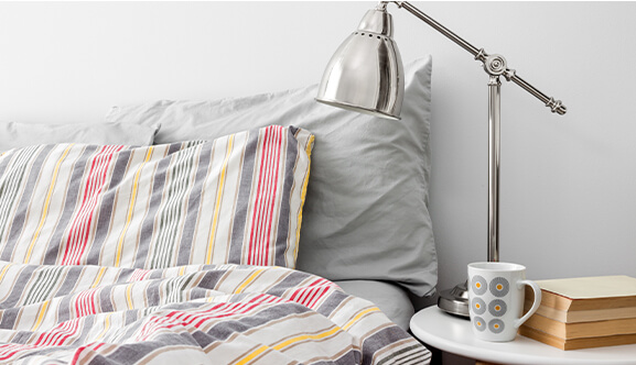 Unmade bed with striped red, grey and yellow bedding with a white bedside table and on top of it a silver metal lamp, white and grey dotted coffee mug and books